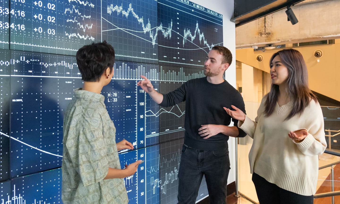 A group of three students in a classroom, focused on a graph on the blackgoard. One student is indicating a point on the graph.