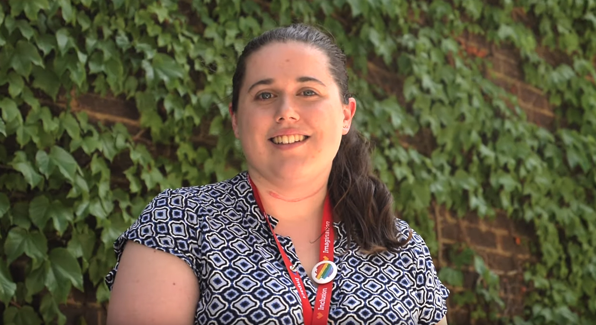 A female wearing an RMIT lanyard, talking to camera, on campus, in front of a leafy green covered building