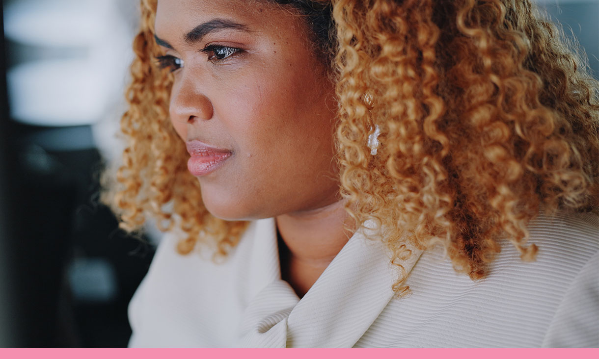 Portrait of a woman in an office. She has strawberry blonde shoulder length curls and wears a white blouse.