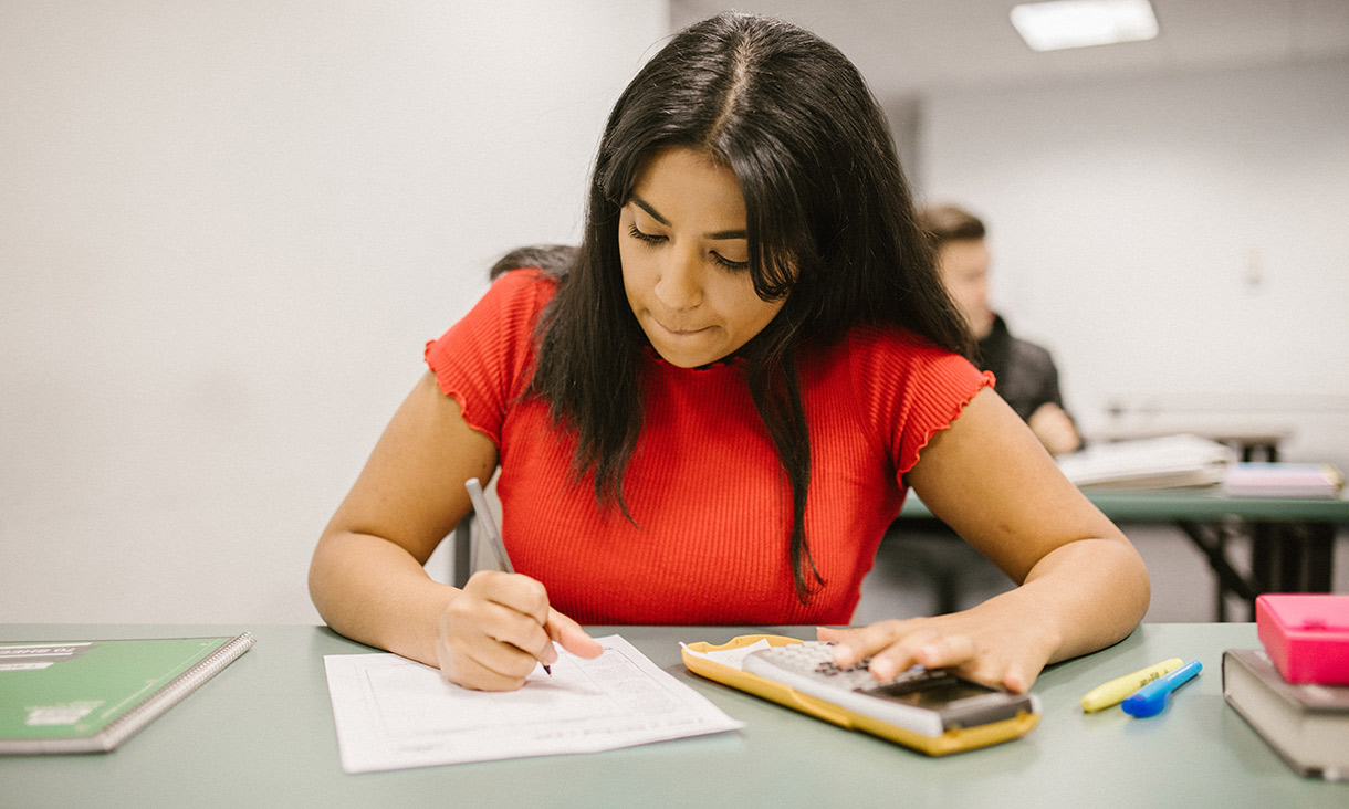 A student working on a desk with a pen, paper and calculator