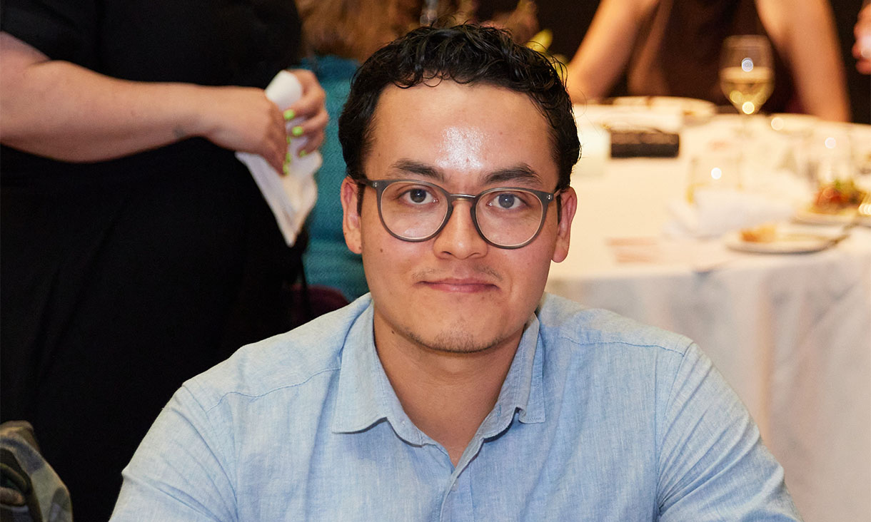 Fahim has short dark hair that curls at the front. He is sitting at a dinner table wearing a light blue shirt and round dark-rimmed glasses.