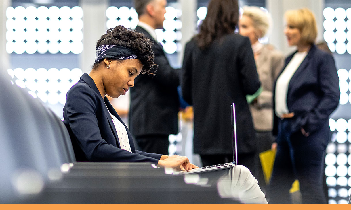 A woman sits in a lecture theatre working on her laptop while a group of people stand around chatting in the background.