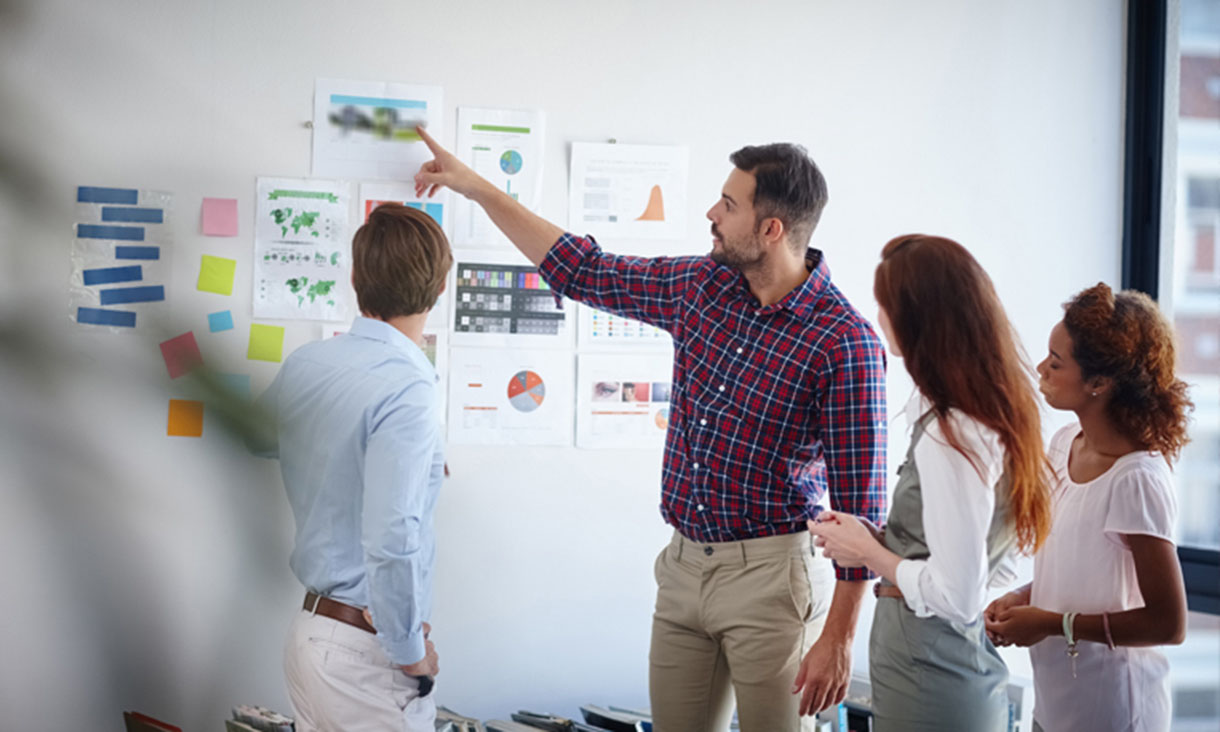 A project manager points to a collection of plans taped to a white wall while his team listens.