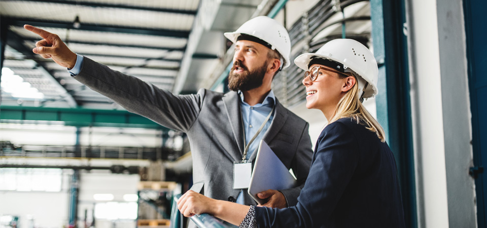 A man wearing a white hard hat and a suit talks to a woman wearing a suit and a white hard hat.