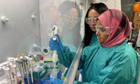 Dr Baoyue Zhang and Dr Sanjida Afrin, wearing lab gowns, gloves and safety glasses, work together at a fume hood while handling laboratory pipettes.
