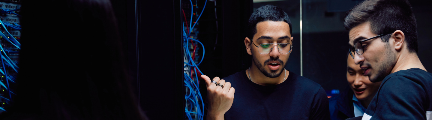 Two students holding laptops standing, looking towards a teacher, all standing in front of very large screens with lines of coloured computer code