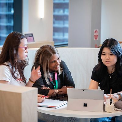 Three students studying at a table, one on a laptop, another smiling, and the third watching.