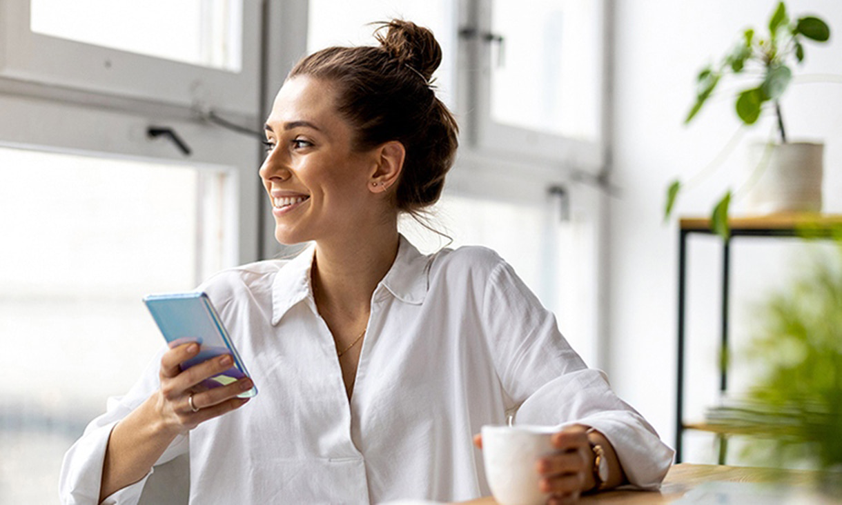 Woman smiling and looking out of window while holding a smart phone and a mug.