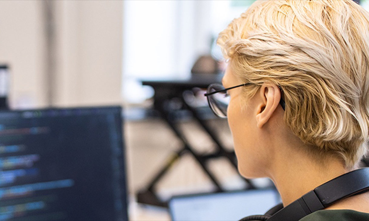 A woman sits at a laptop that has code on the screen with headphones around her neck.