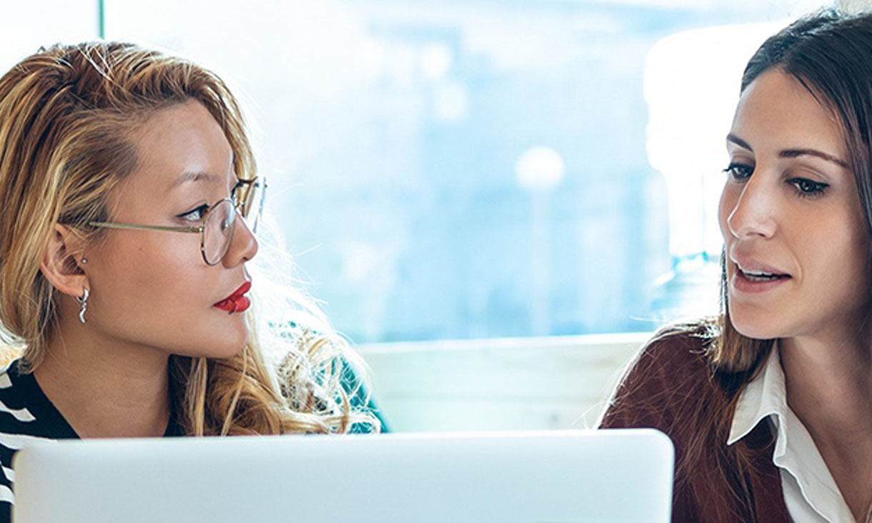Two women have a discussion while looking at a laptop screen.