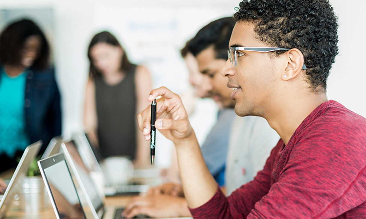 A man attends a meeting with others at a shared desk with laptops.